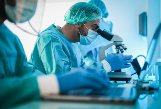 Researchers in full protective gear working in a laboratory, one using a microscope, another on a laptop.